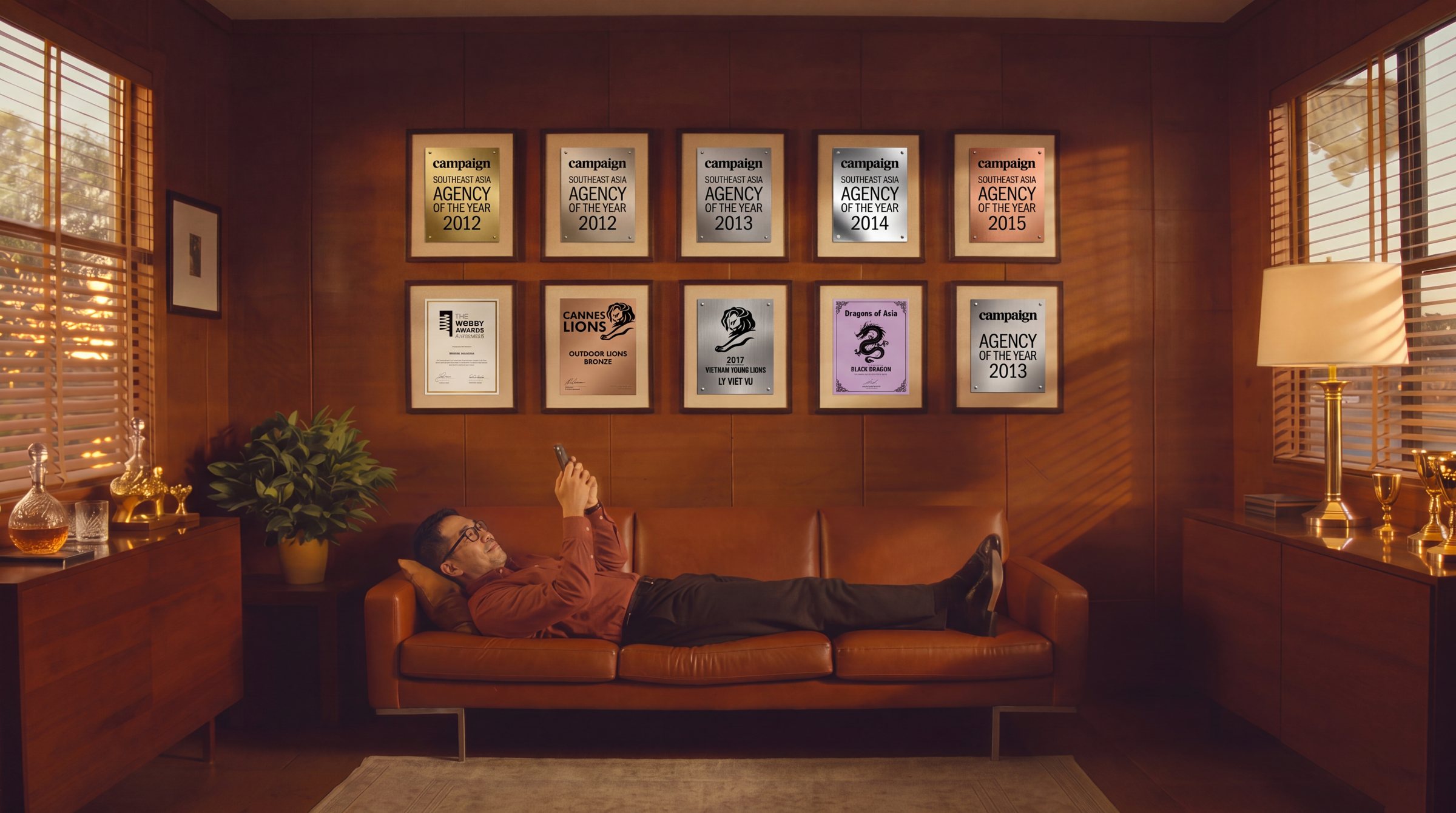 Ly lying on a leather sofa in a 1960s office under ten framed agency awards
